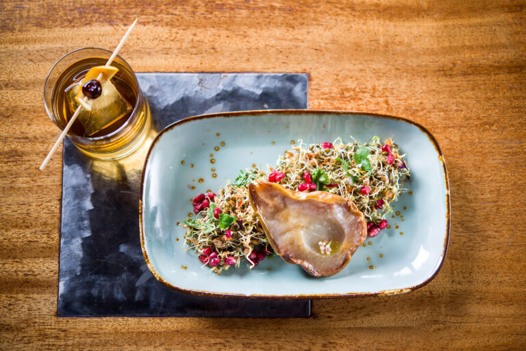 A roasted pear sitting on a bed of sprouts and pomegranate with a handcrafted old fashioned sitting next to it.