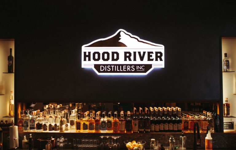 A neon Hood River Distillers sign with bottles of house made liquor on the shelves.