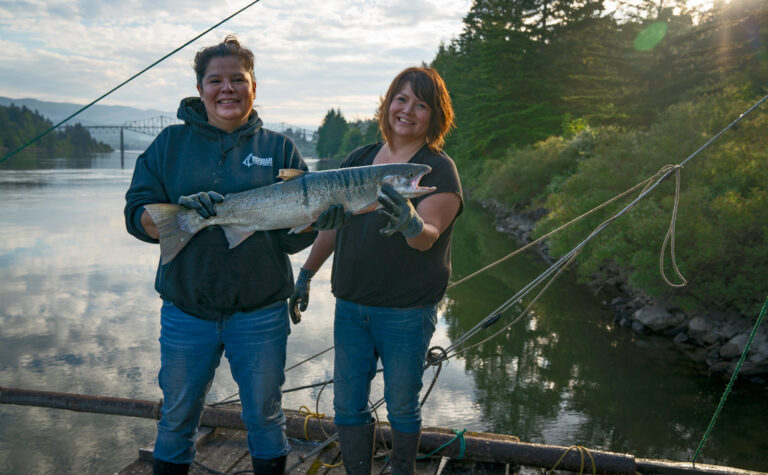 Two people holding a salmon in front of the river.